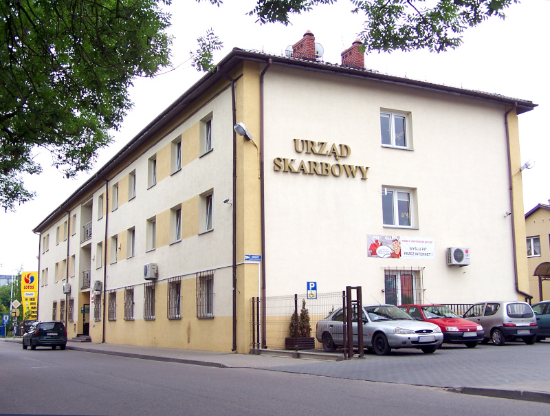 Budynek Urzędu Skarbowego w Płońsku. Wzdłuż budynku biegnie droga asfaltowa, na której zaparkowany jest samochód. Przy budynku parking, a na nim kilka samochodów.