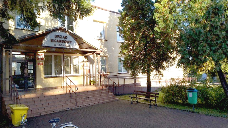 Budynek Urzędu Skarbowego w Łosicach. Przed budynkiem chodnik, drzewa, ławka.