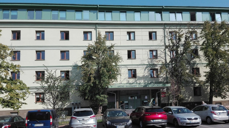 Budynek Urzędu Skarbowego Warszawa-Targówek. Przed budynkiem drzewa, parking i kilka samochodów.