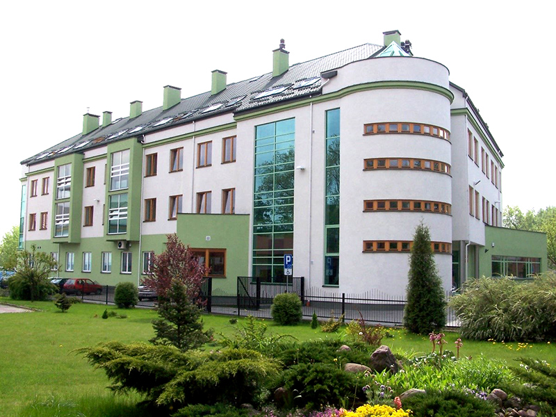    Budynek Urzędu Skarbowego w Mińsku Mazowieckim. Przed budynkiem parking i trawnik, a na nim dużo drzew i krzewów iglastych.