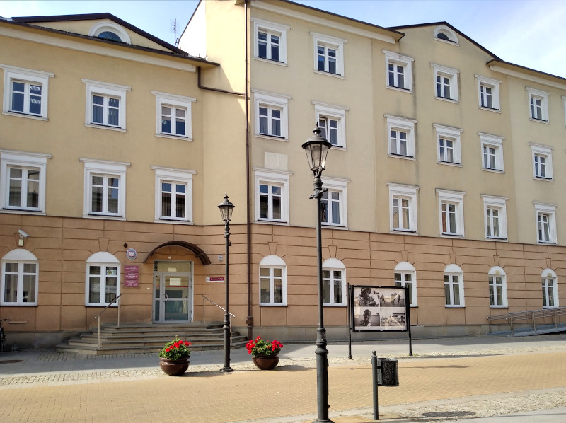 Budynek Urzędu Skarbowego w Ciechanowie. Przed budynkiem gablota ze zdjęciami, latarnie, ulica. 