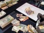 Funkcjonariusz liczy znalezione u podejrzanych pieniądze. Na biurku pliki banknotów w różnych walutach.
