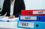 Na pierwszym planie segregatory z napisem VAT, w tle osoba, która przegląda dokumenty.