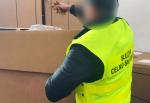 Funkcjonariusz służby celno-skarbowej w żółtej kamizelce trzyma w ręku suszony tytoń nad dużym kartonowym pudłem.