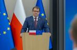 Szef KAS Marcin Łoboda przy mównicy z mikrofonami, za nią widoczne są flagi Polski i Unii Europejskiej.