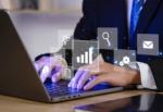 Dłonie na klawiaturze laptopa z ikonami symbolizującymi wyszukiwanie, wzrost wykresu, ustawienia i komunikację, sugerujące pracę z danymi i zarządzanie informacjami.