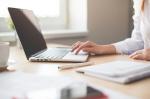 Fotografia przedstawia osobę pracującą przy laptopie na jasnym biurku z notesem, długopisem i filiżanką kawy.