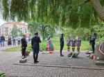 Uroczystość z udziałem umundurowanych żołnierzy i harcerek przy pomniku z kwiatami i zniczami, pod drzewem.