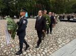 Uroczysta ceremonia składania wieńców przez osoby w mundurach i garniturach na placu z kamienną nawierzchnią, z obecnością publiczności i flag państwowych w tle.