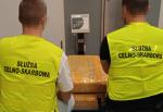 Fotografia przedstawia dwóch funkcjonariuszy Służby Celno-Skarbowej w odblaskowych żółtych kamizelkach, stojących przy zabezpieczonej taśmą paczce.
