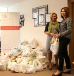 Pani Dyrektor Teresa Warszawska oraz Rzecznik IAS w Warszawie Anna Szczepańska trzymają torbę z darami. Na drugim planie ułożone  wypełnione 